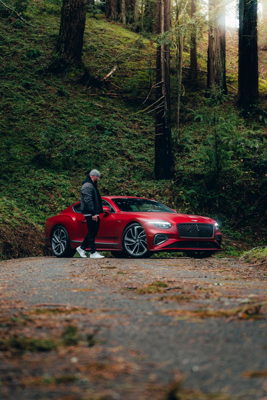Max beside a red Bentley Continental GT on a redwood road