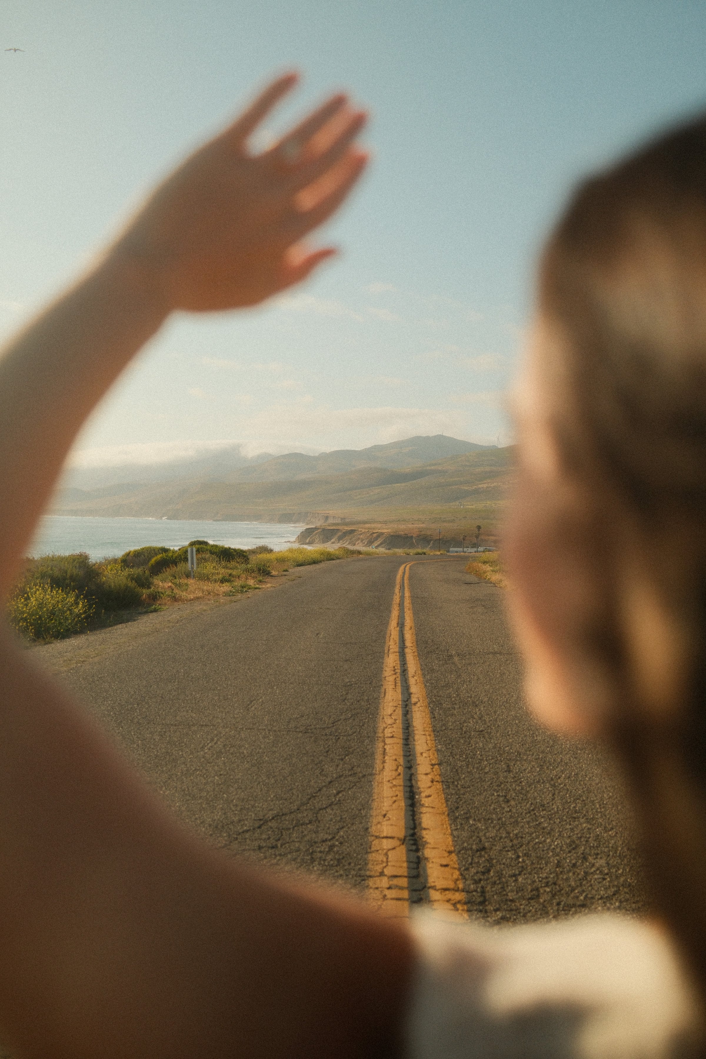 A hand reaching out of a car window over a golden-hour coastal road