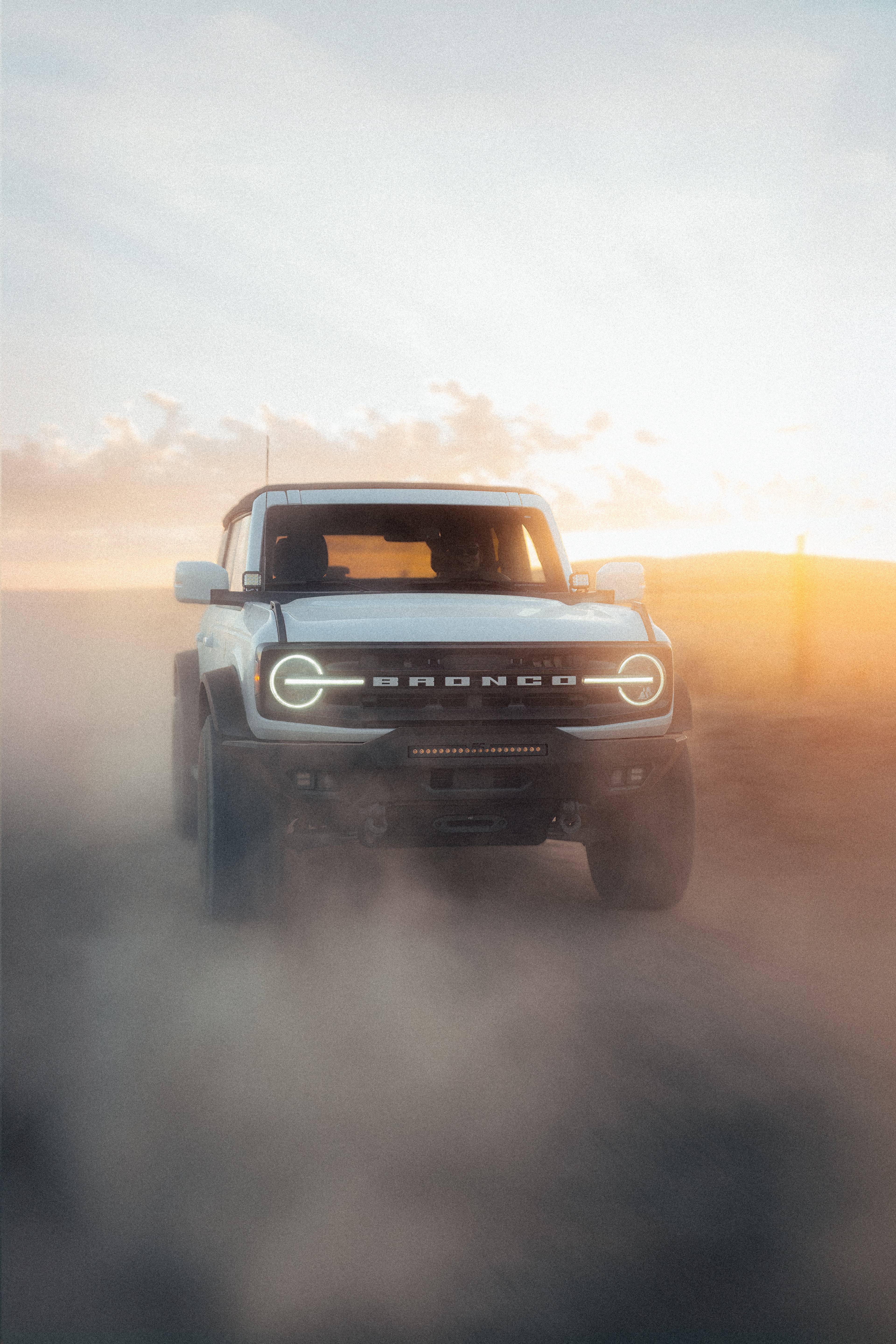 Ford Bronco emerging from heavy dust at sunset, grille lights cutting through