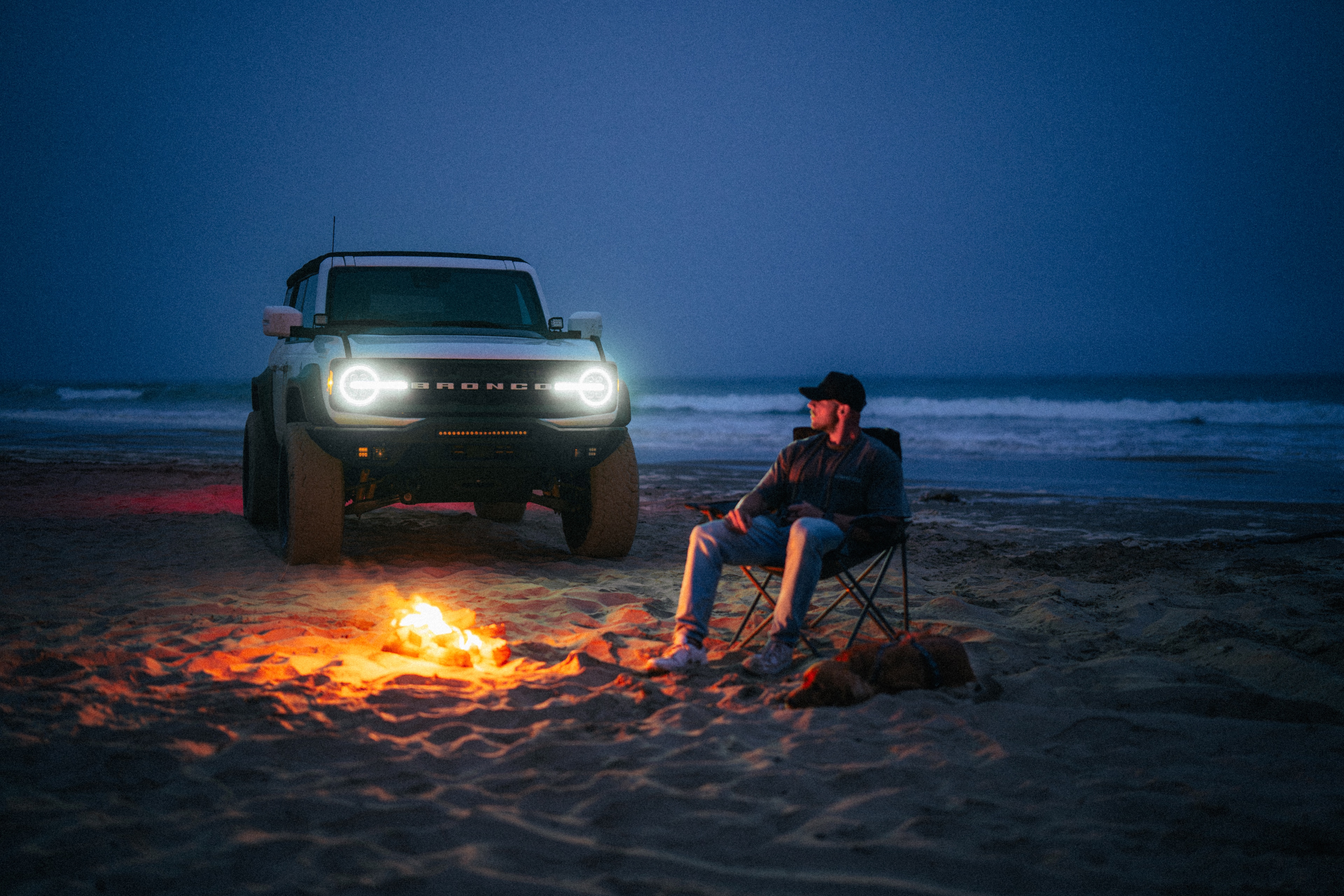 Ford Bronco parked on the beach at blue hour, headlights lit, a campfire burning beside a figure in a camp chair