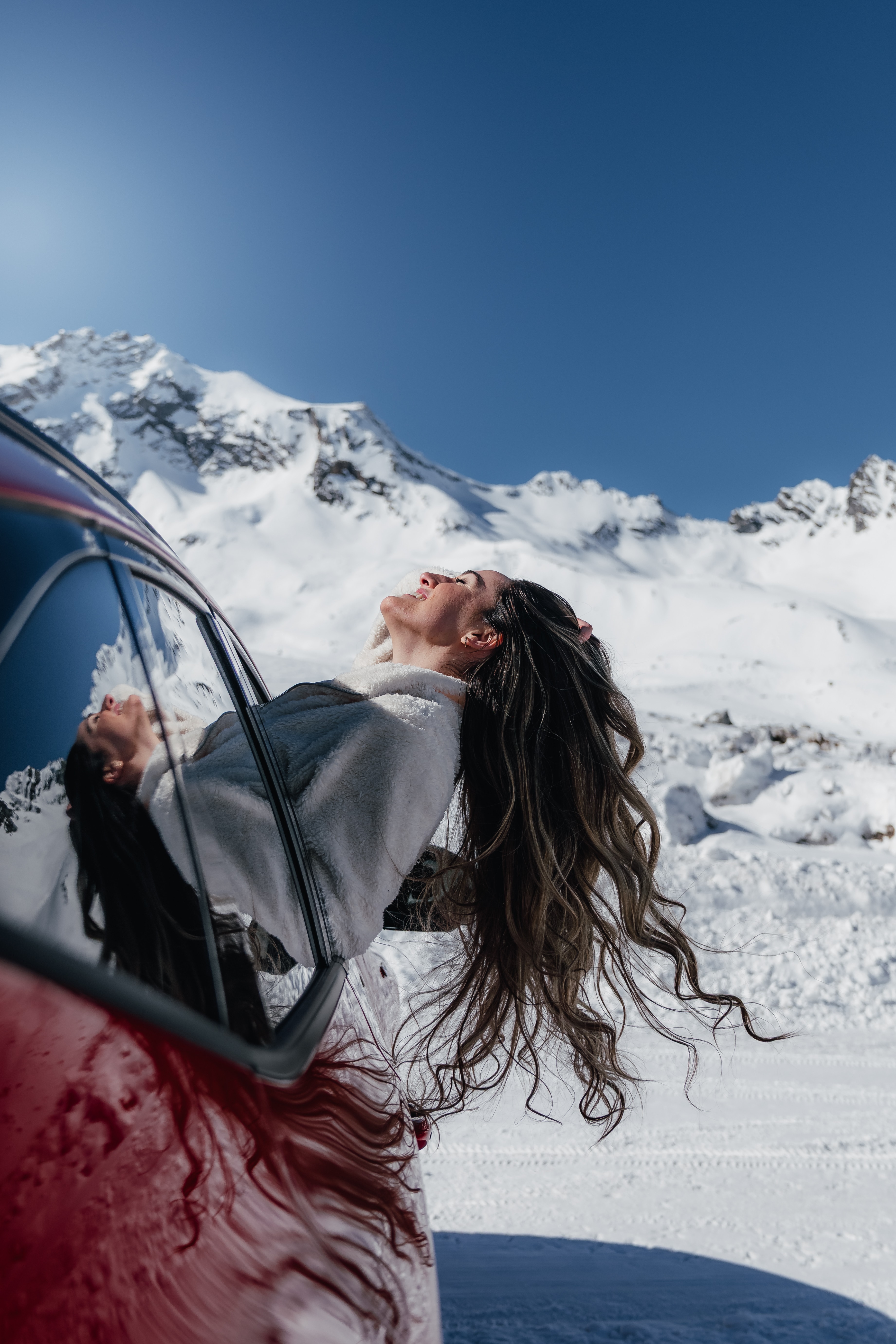 A passenger leans out of the red Hyundai into the sun, alpine peaks behind