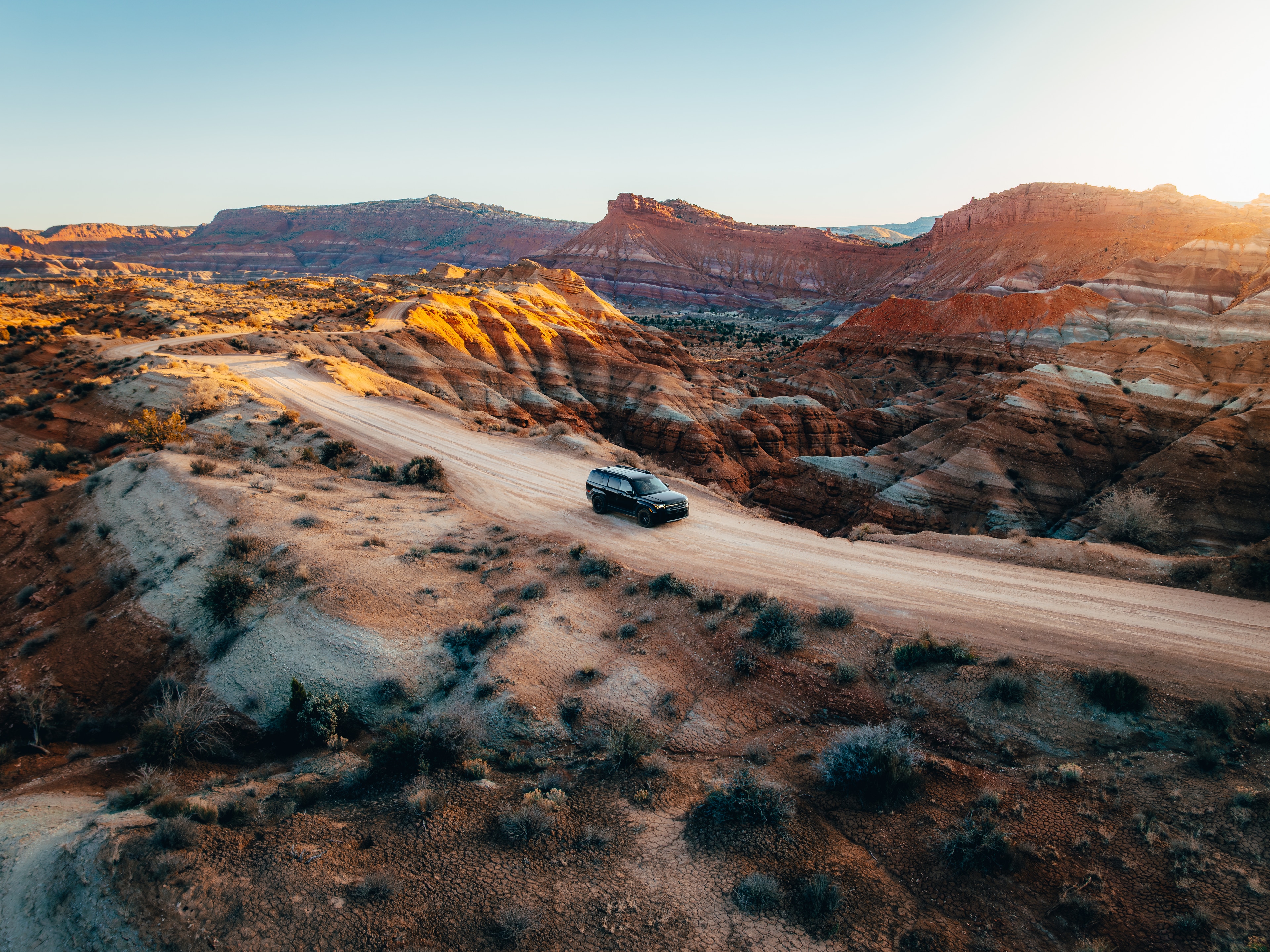 Hyundai Santa Fe on a winding dirt road descending through banded red rock badlands at sunset, Utah