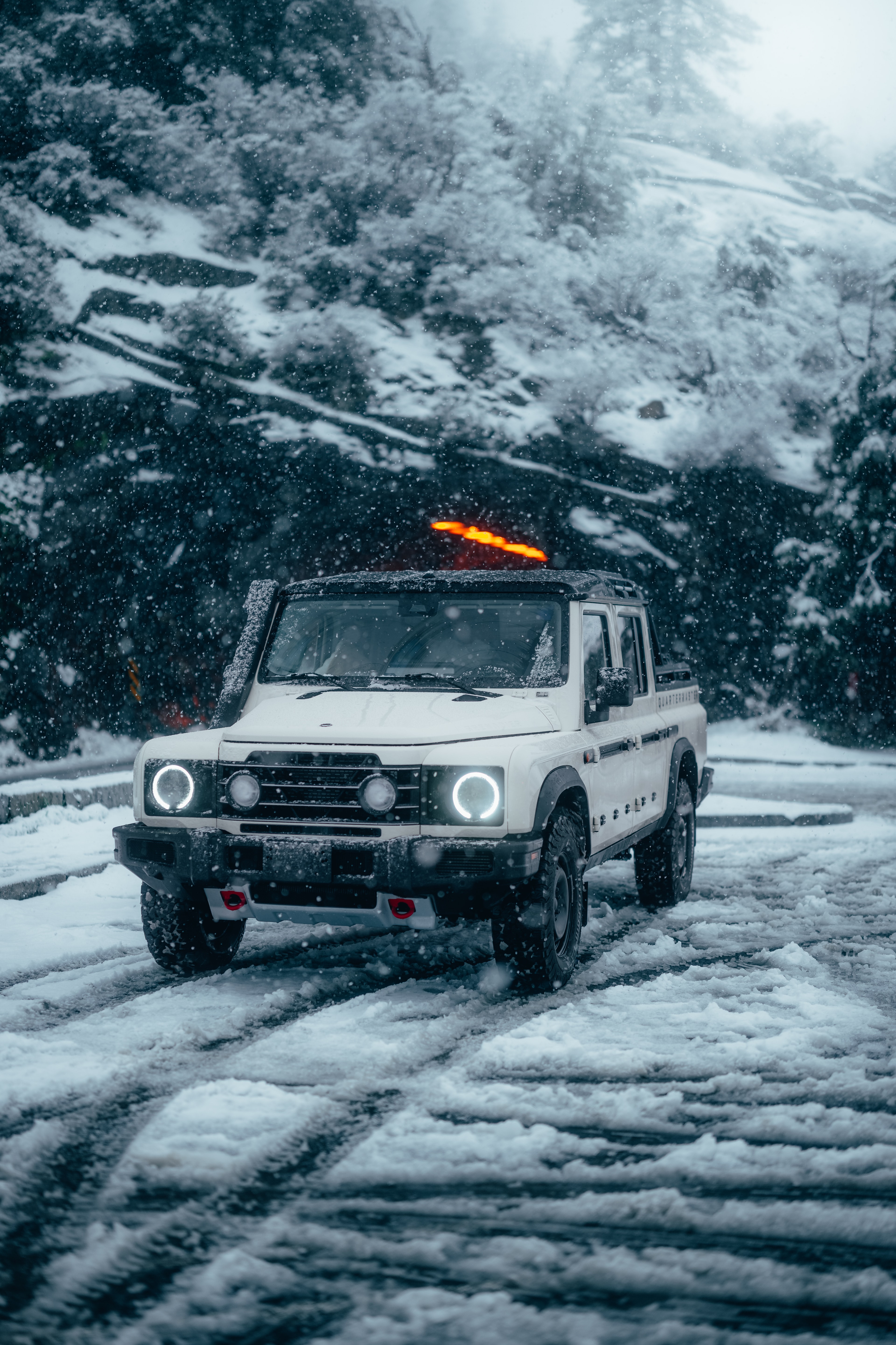 INEOS Grenadier Quartermaster front three-quarter in heavy falling snow, round headlights glowing