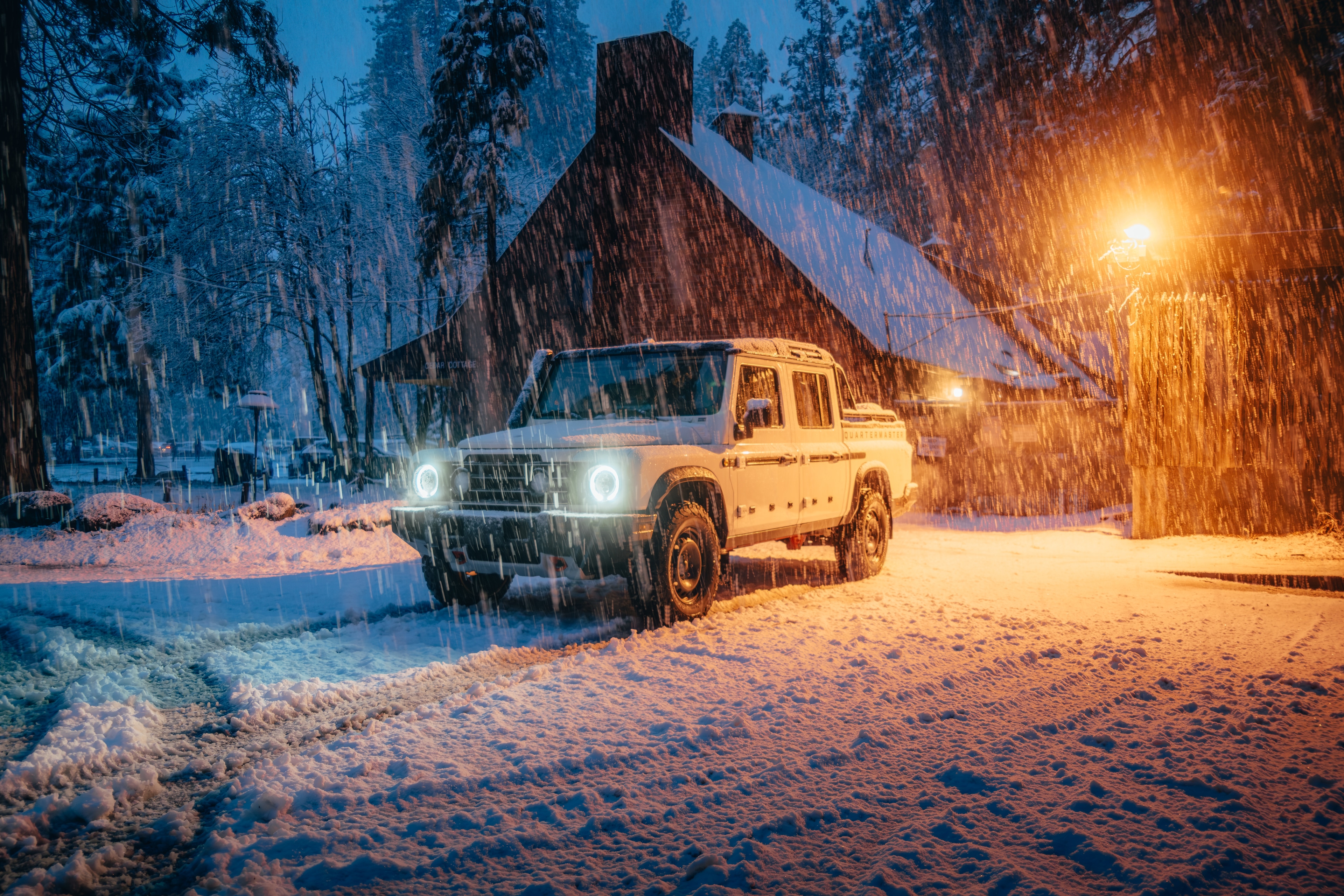 INEOS Grenadier Quartermaster at blue hour beside a snowy A-frame cabin, headlights cutting through heavy snow