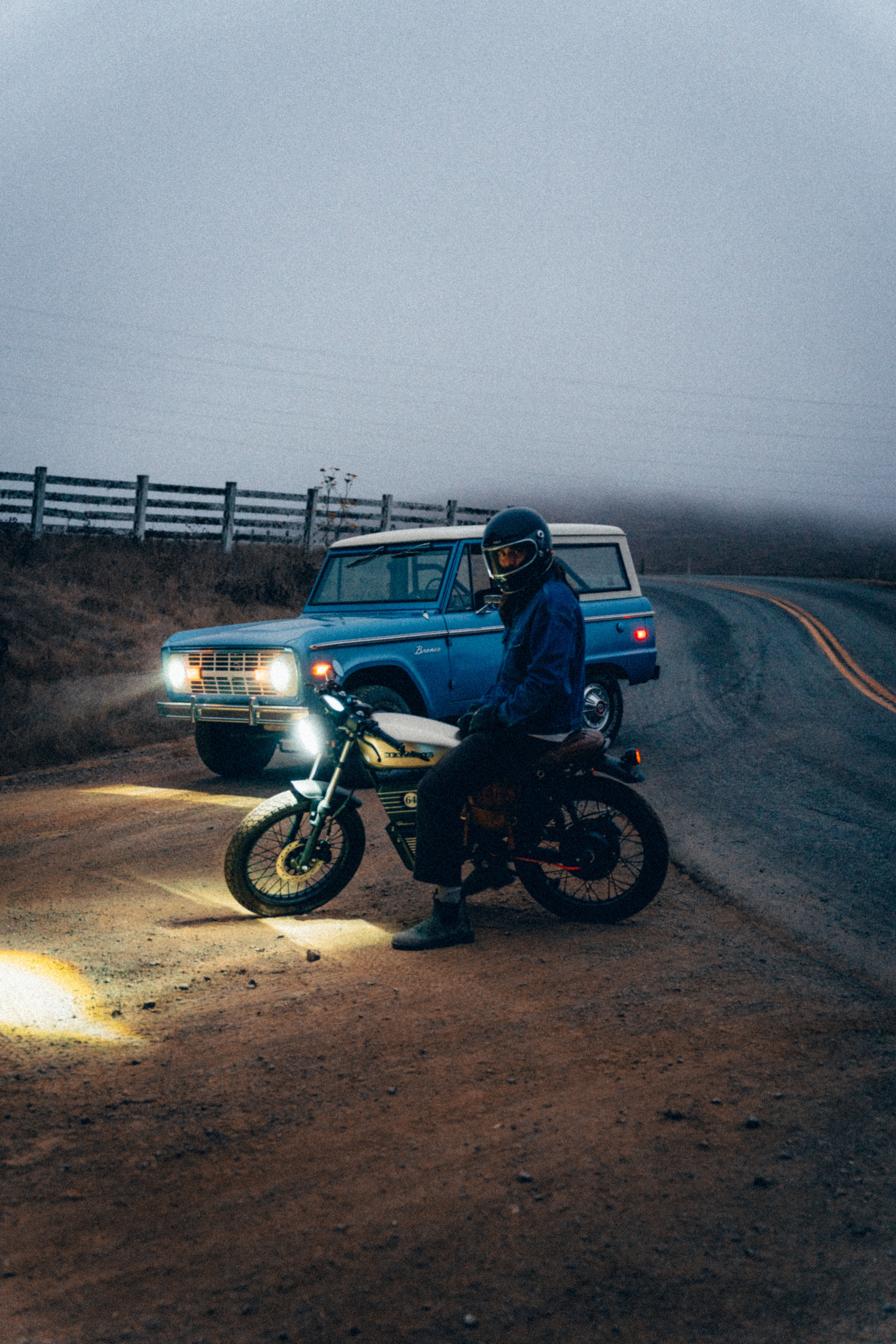Rider stopped on the Beachman at dusk beside a vintage blue Ford Bronco, headlights on