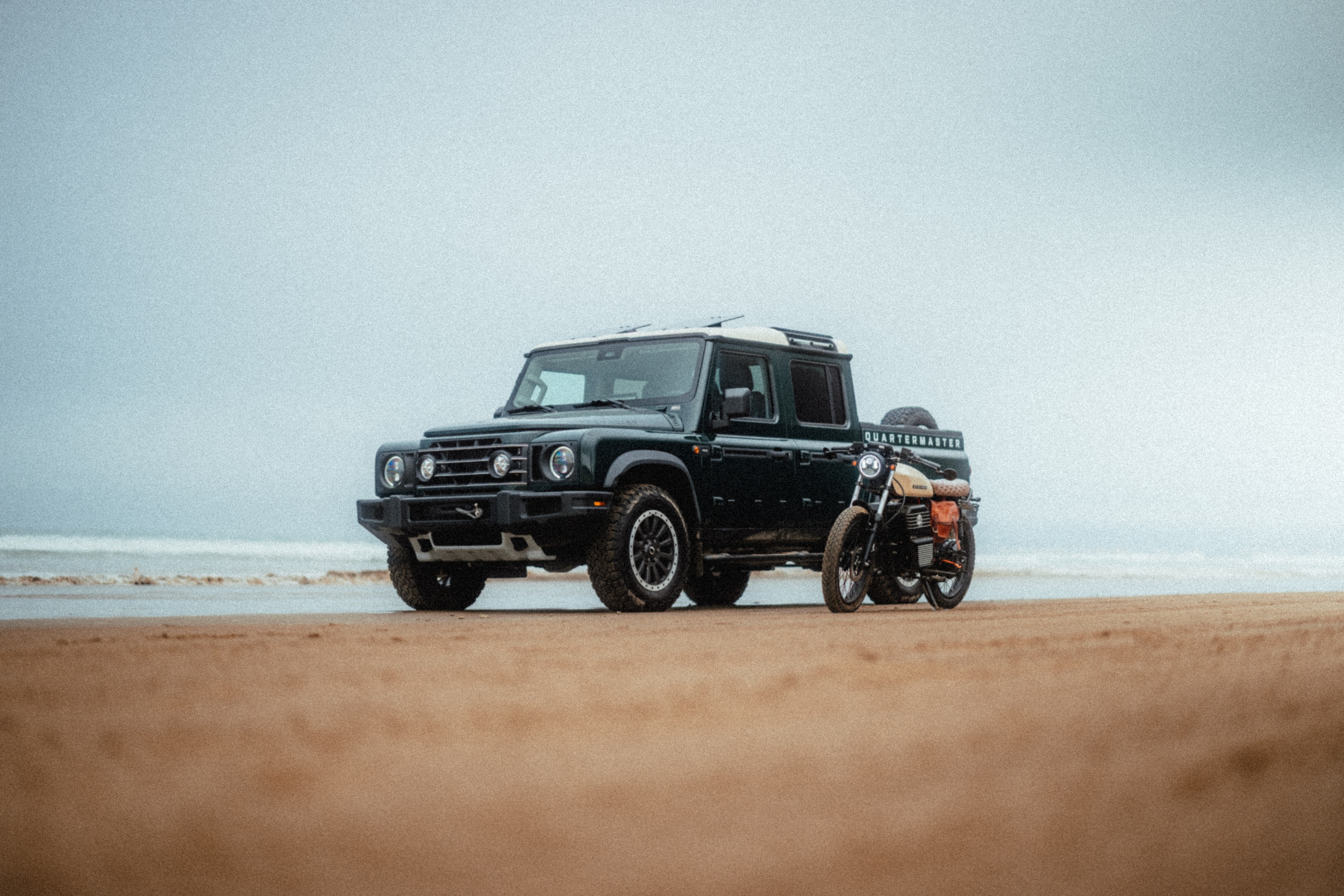 Beachman cafe racer parked beside a black INEOS Grenadier Quartermaster on the beach