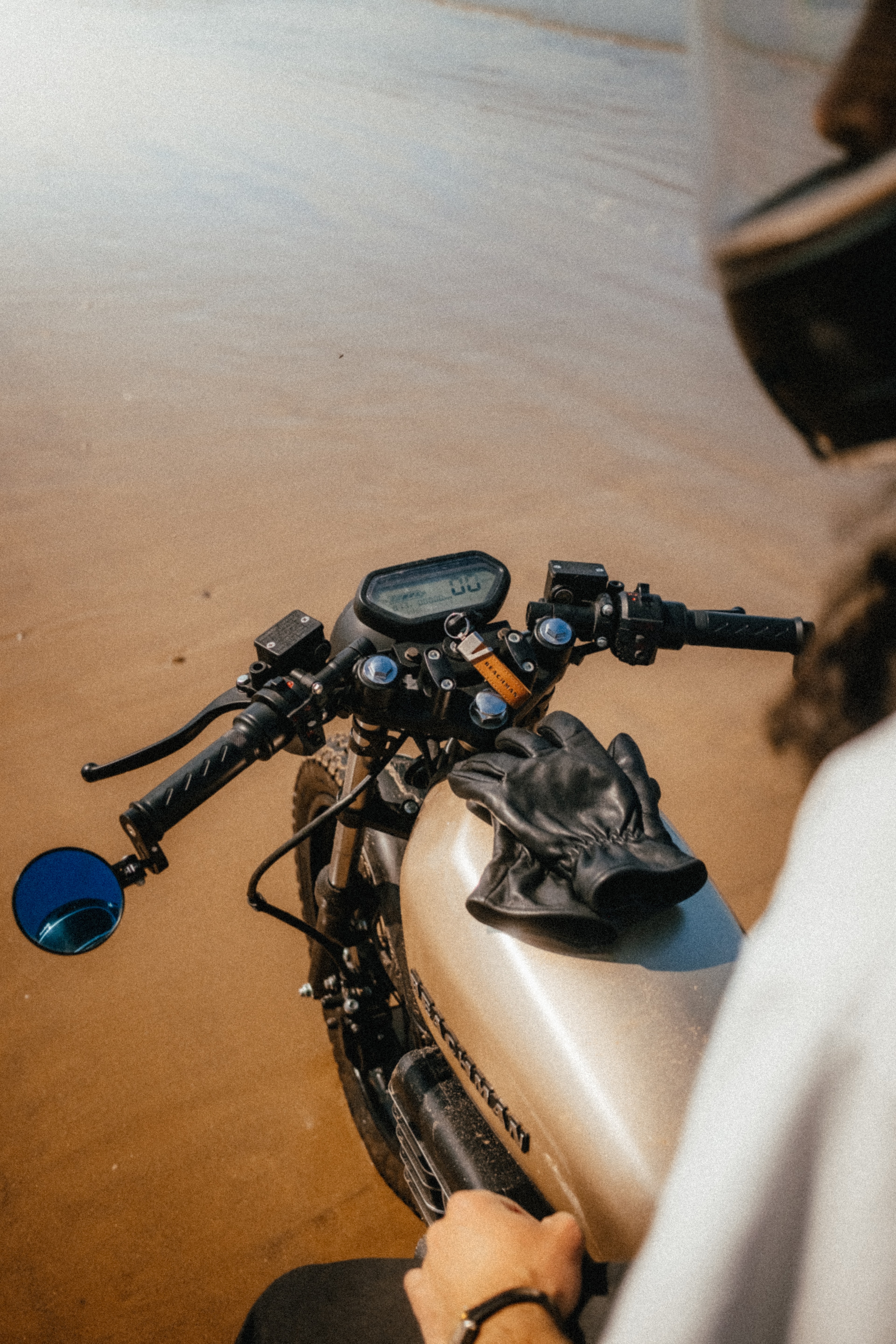 Rider's-eye view over the Beachman handlebars and gauge, gloves on the tank, sand below