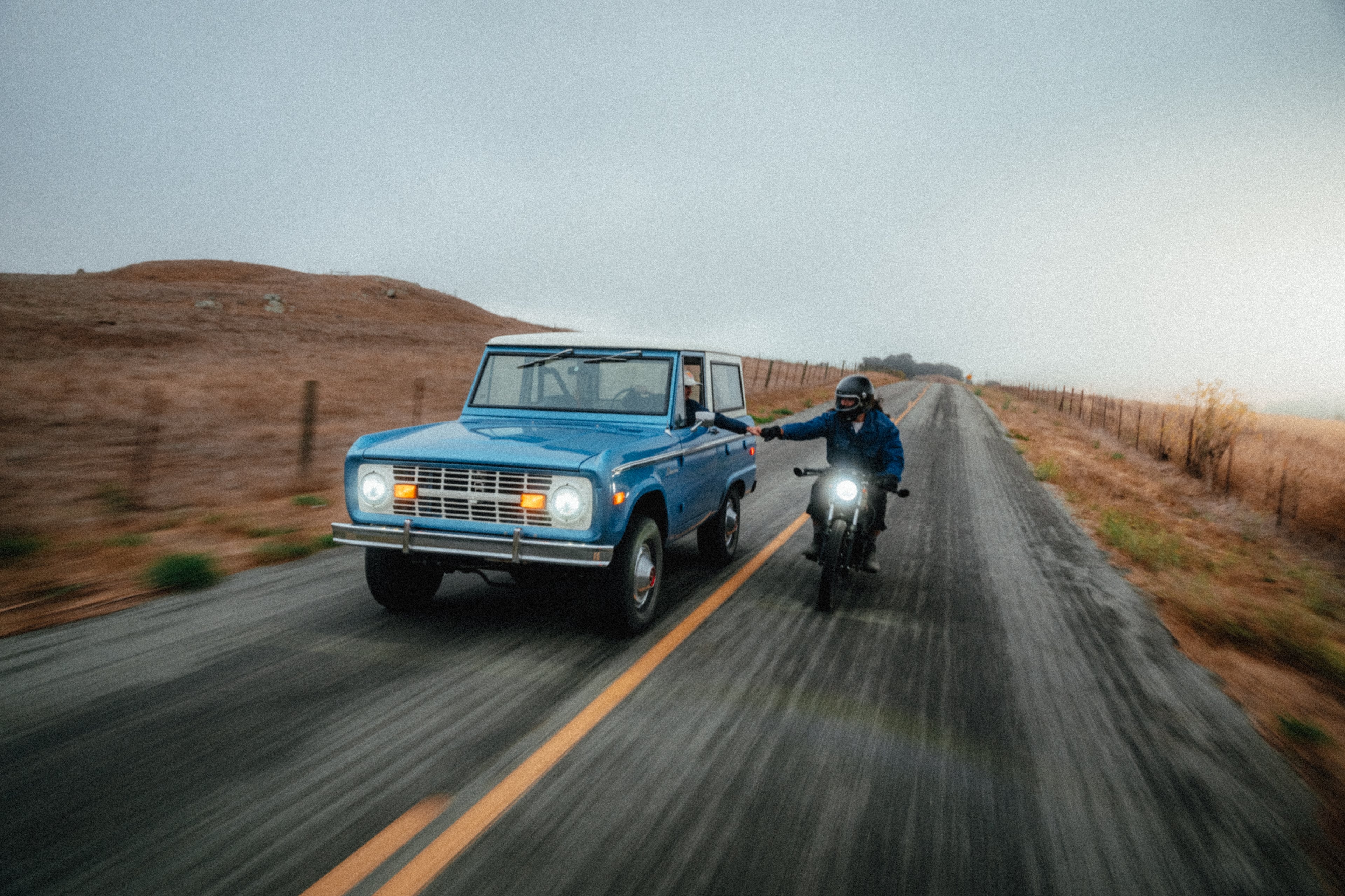 Rider on the Beachman cafe racer alongside a vintage blue Ford Bronco on a foggy Central Coast road, in motion