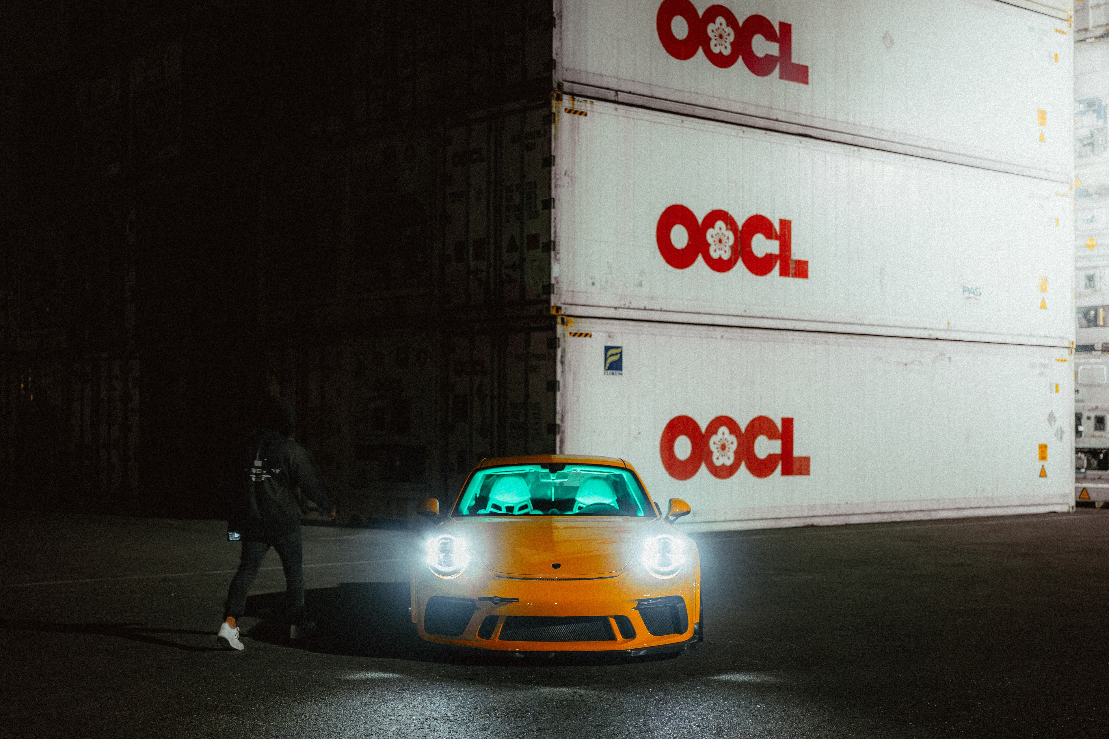Yellow Porsche 911 GT3 at night against shipping containers, headlights blazing