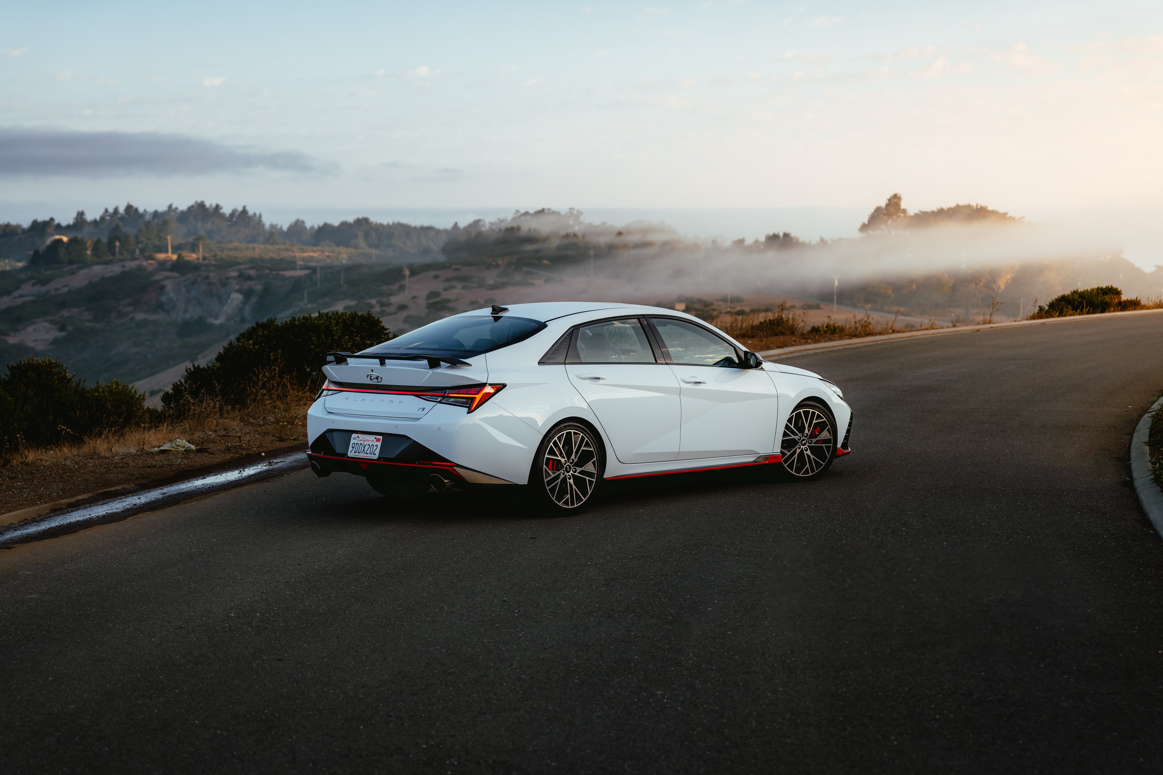 Hyundai Elantra N rear three-quarter at a coastal overlook, fog rolling over the forested hills at golden hour