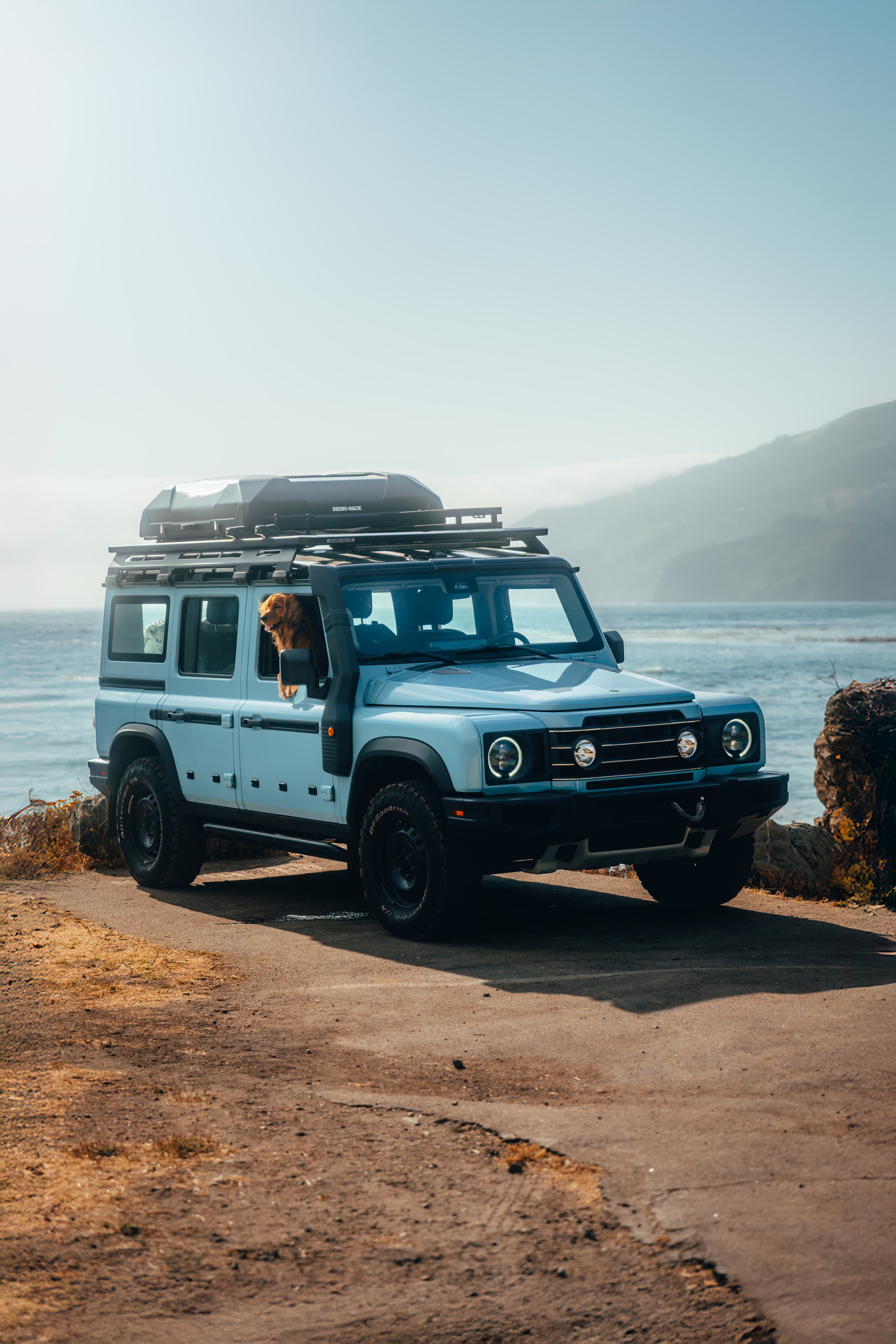 Light blue INEOS Grenadier at a Big Sur coastal pullout, a dog looking out the window