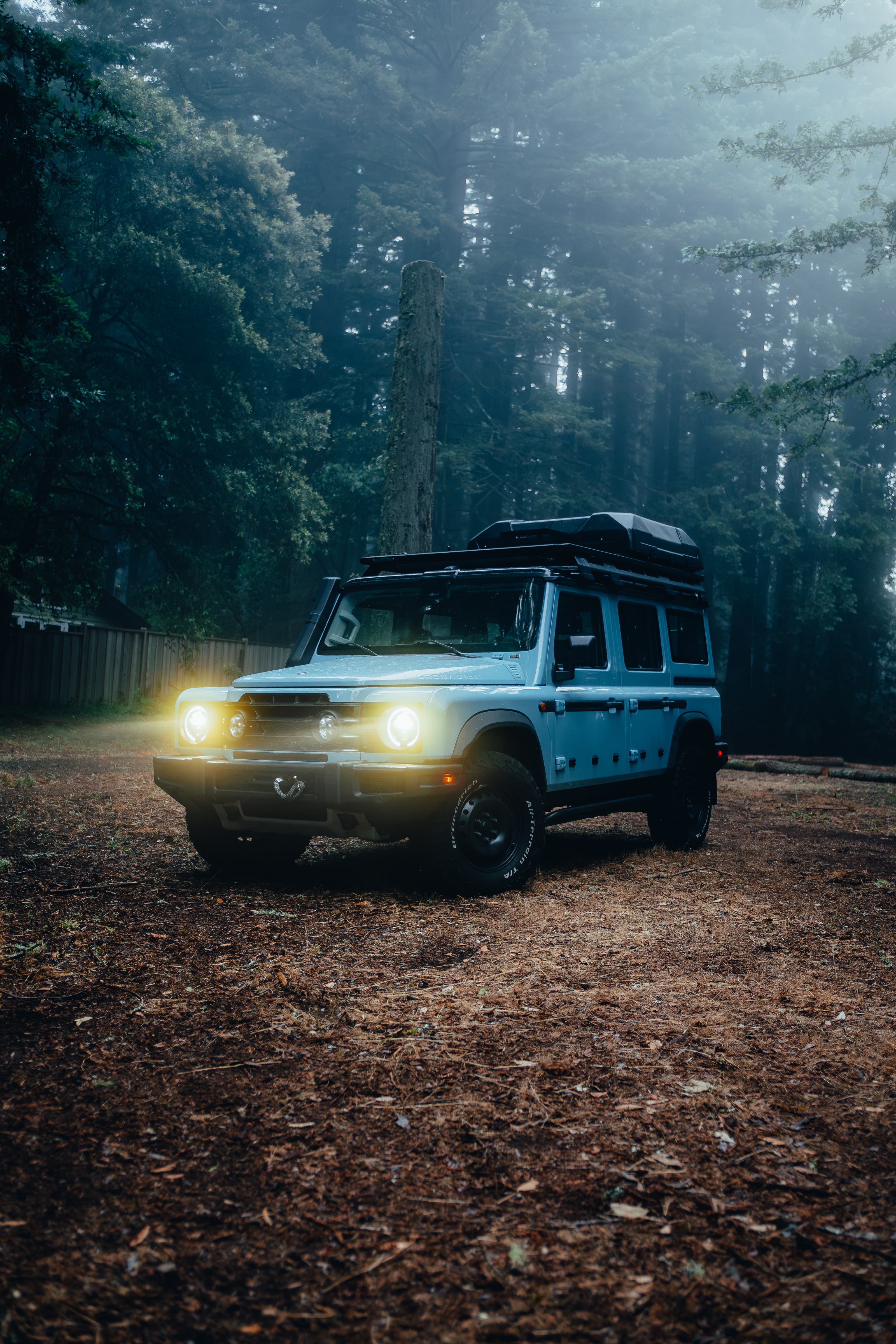 Light blue INEOS Grenadier in a dark redwood clearing, headlights on
