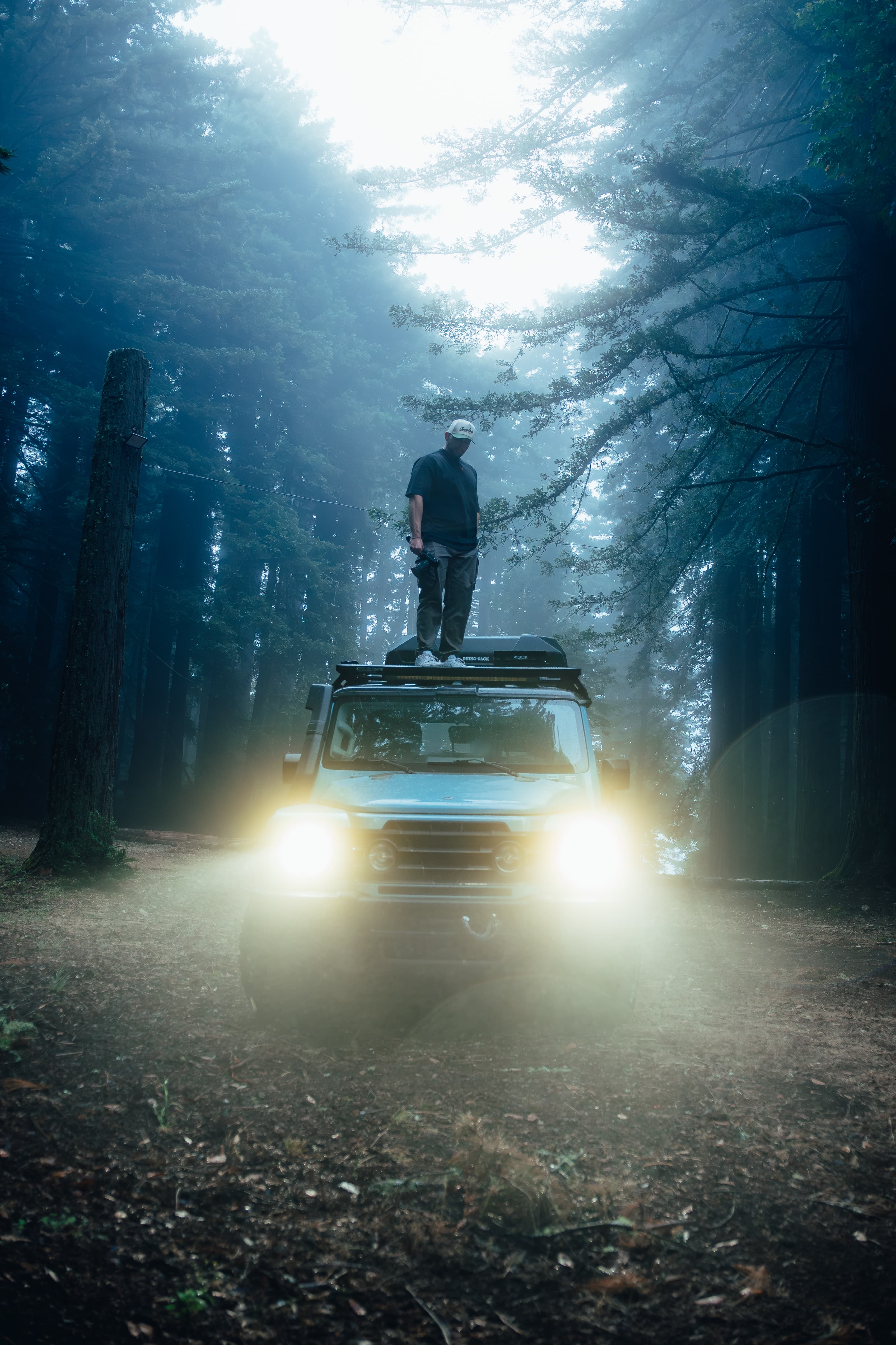 A person standing on the roof of the INEOS Grenadier in a foggy forest, headlights below