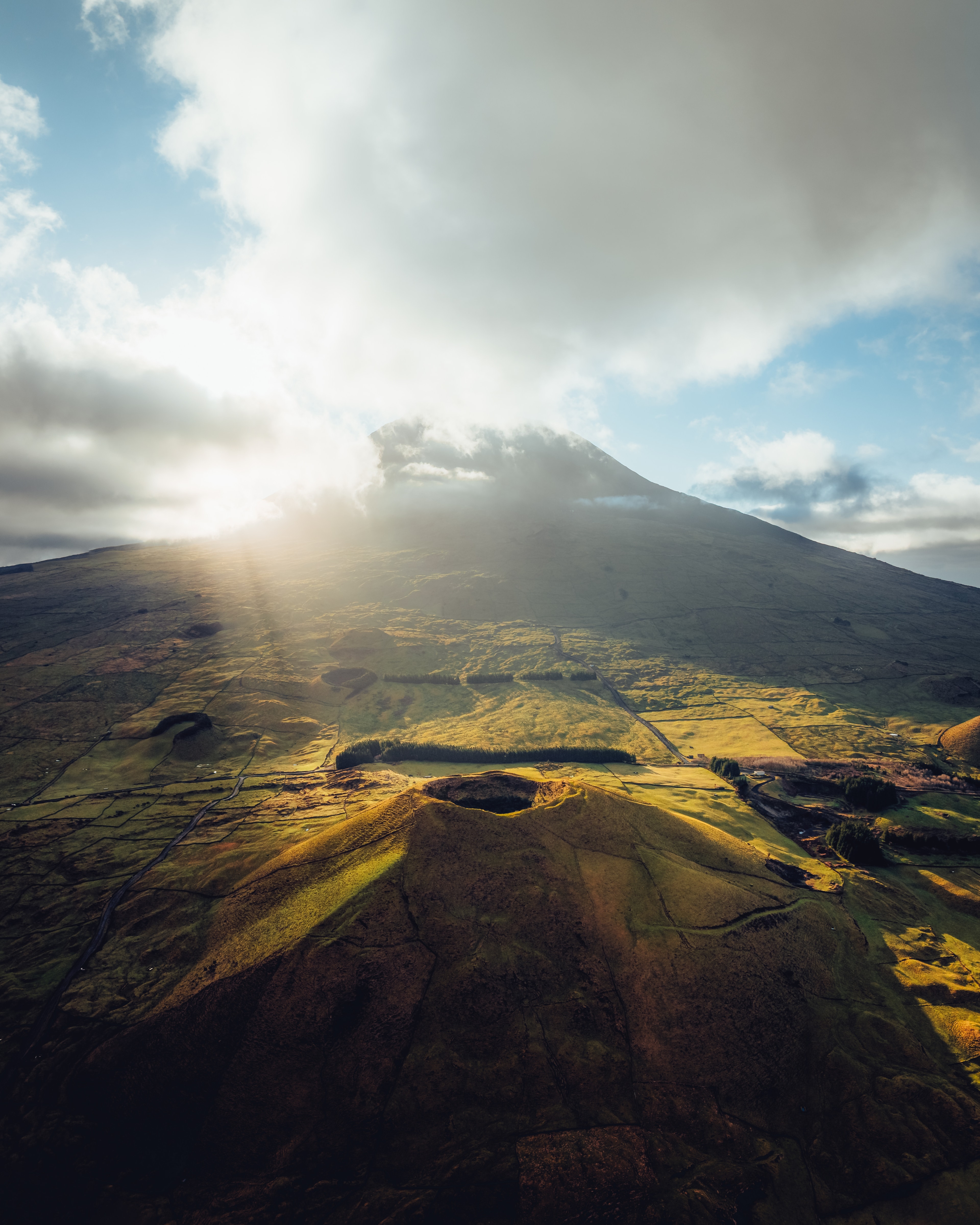 A road curving down into a vast green volcanic caldera, sun flaring over the rim
