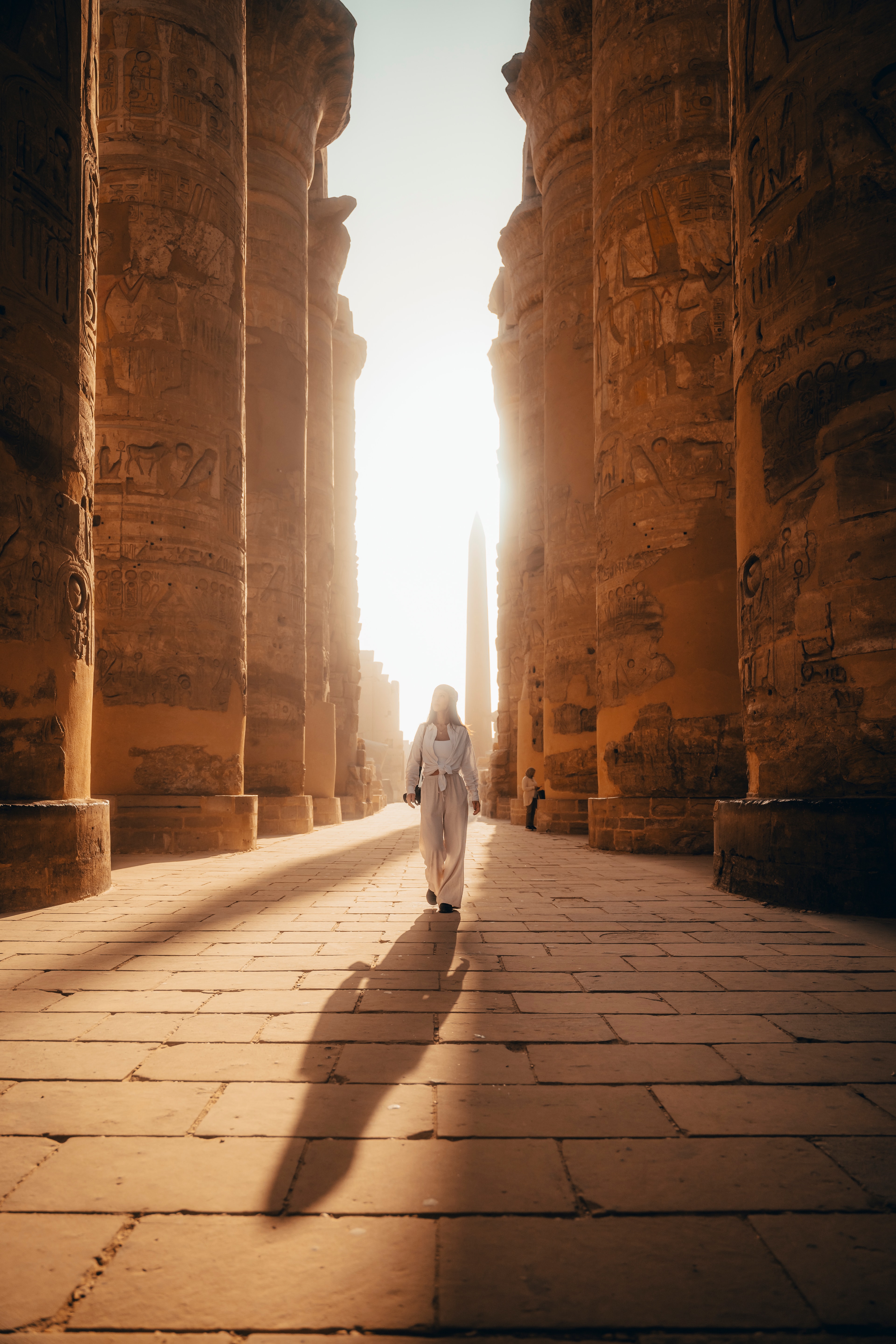 A figure in white walking the central aisle of the Karnak hypostyle hall, sun ahead and a long shadow behind
