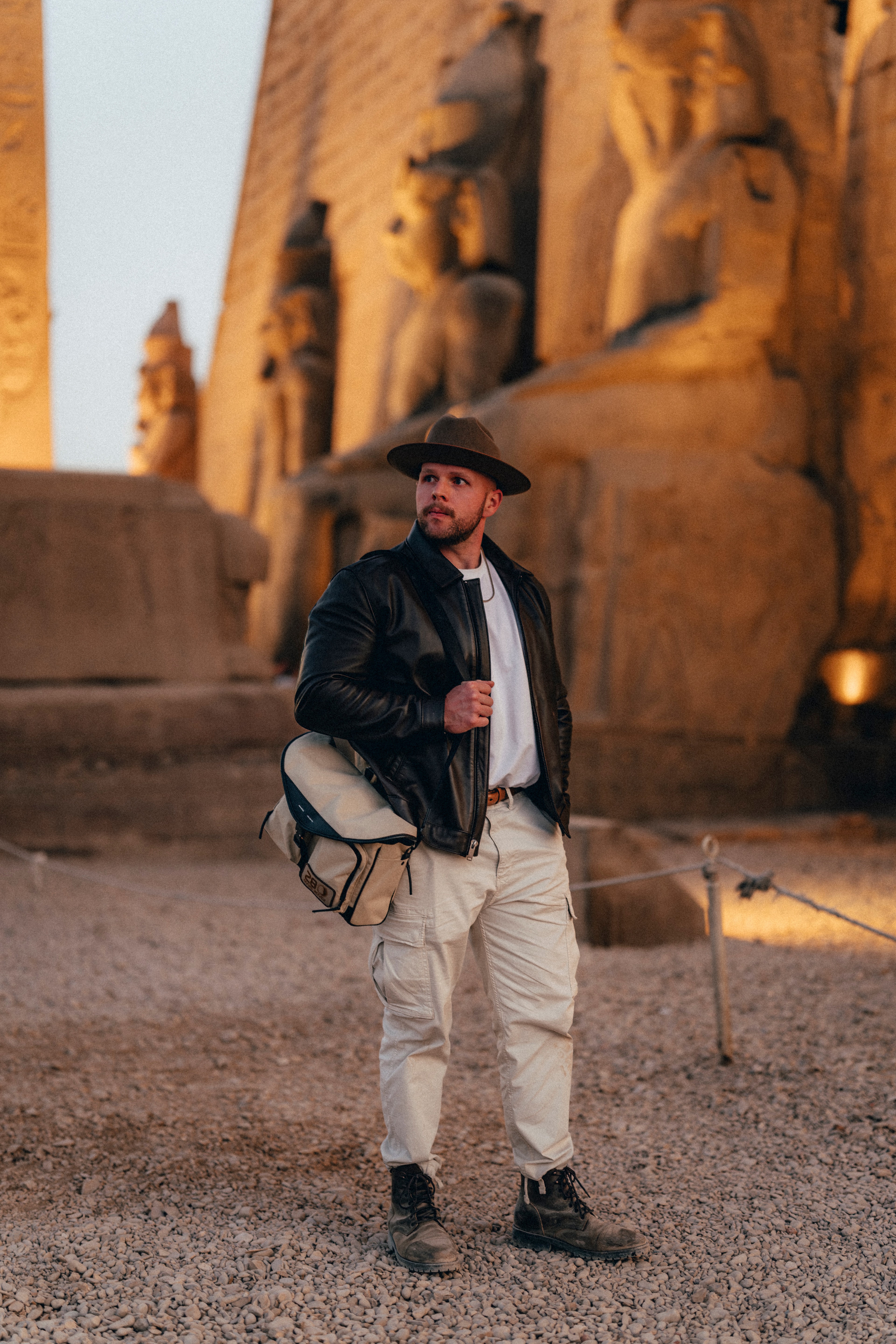 A man in a hat and leather jacket at the entrance to Luxor temple at golden hour, statues of the pharaohs behind him