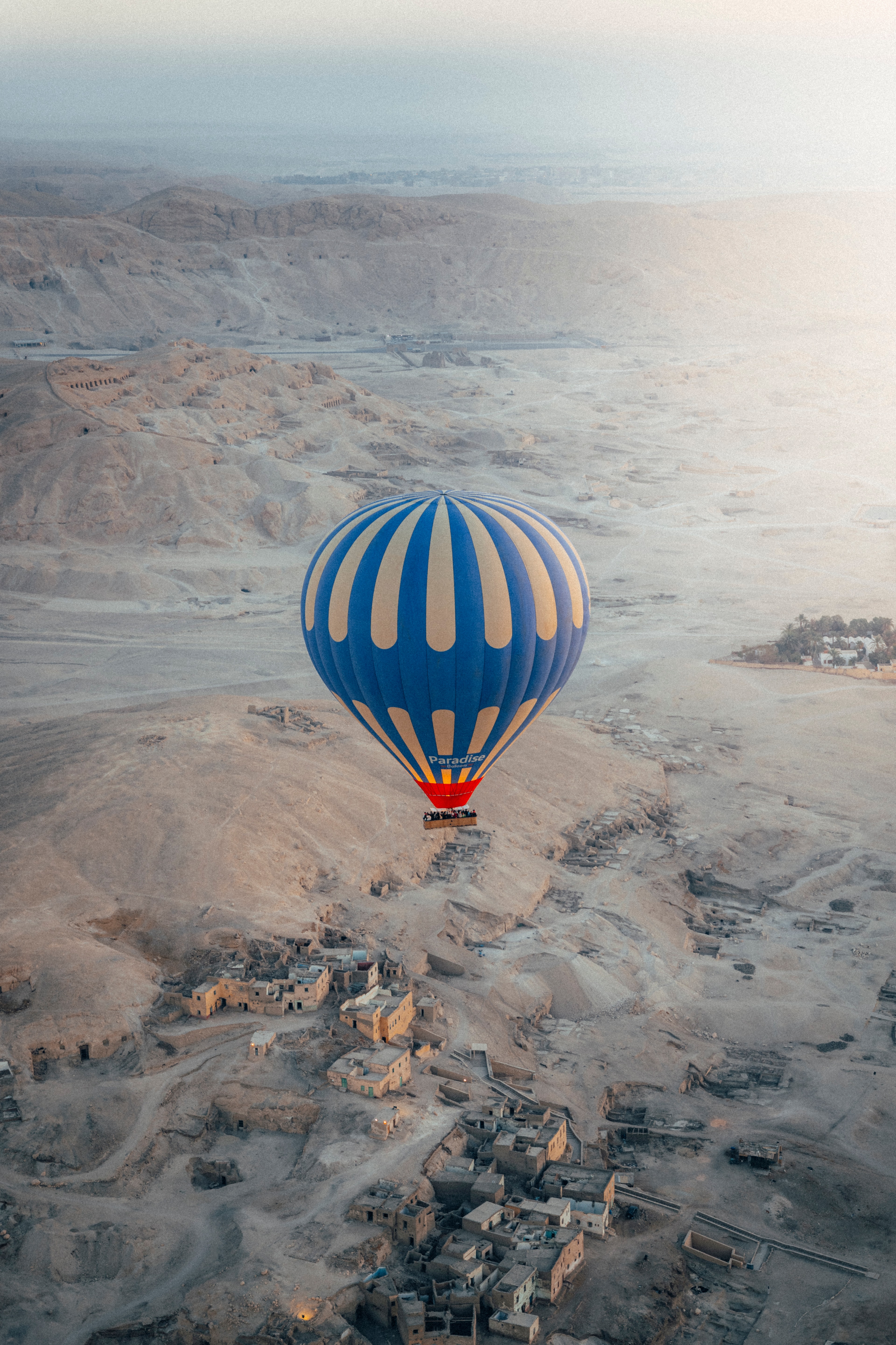 A blue-and-cream hot air balloon drifting over the Luxor west bank village at sunrise