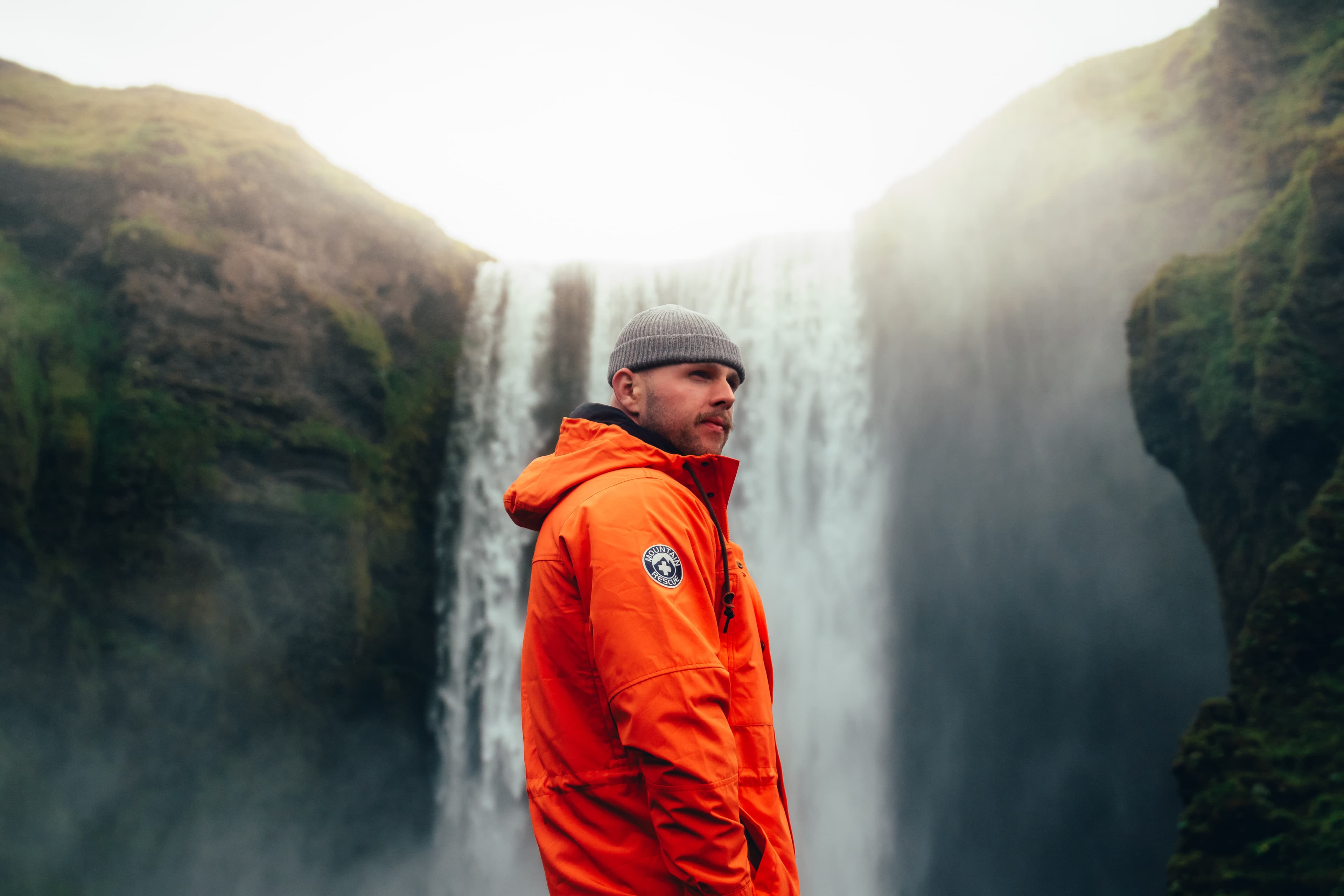 A man in an orange Alpine Design shell jacket and beanie before a misty Icelandic waterfall