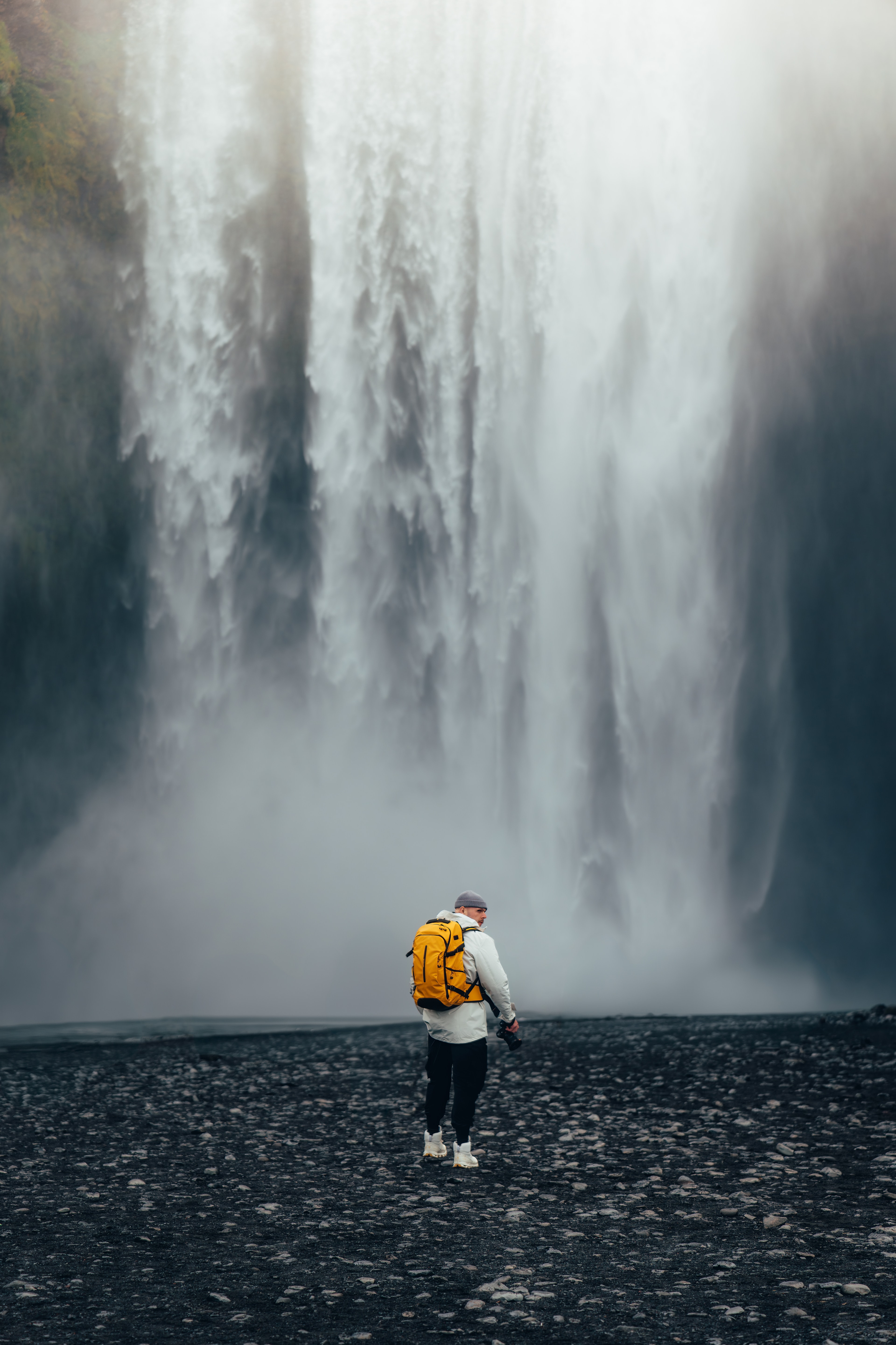 A traveler with a yellow pack dwarfed by a tall curtain of falling water