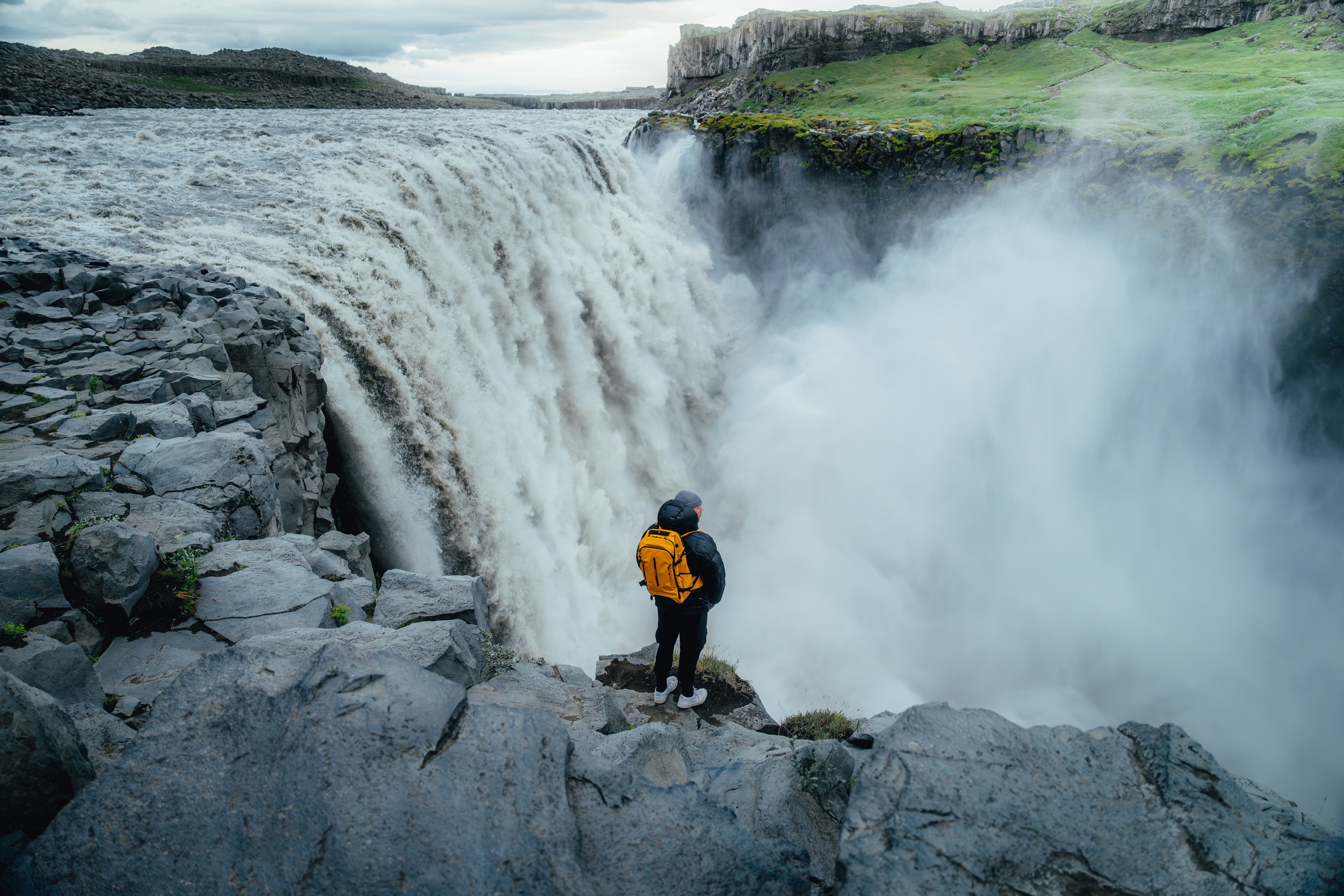 A traveler with a yellow pack at the thundering edge of Dettifoss