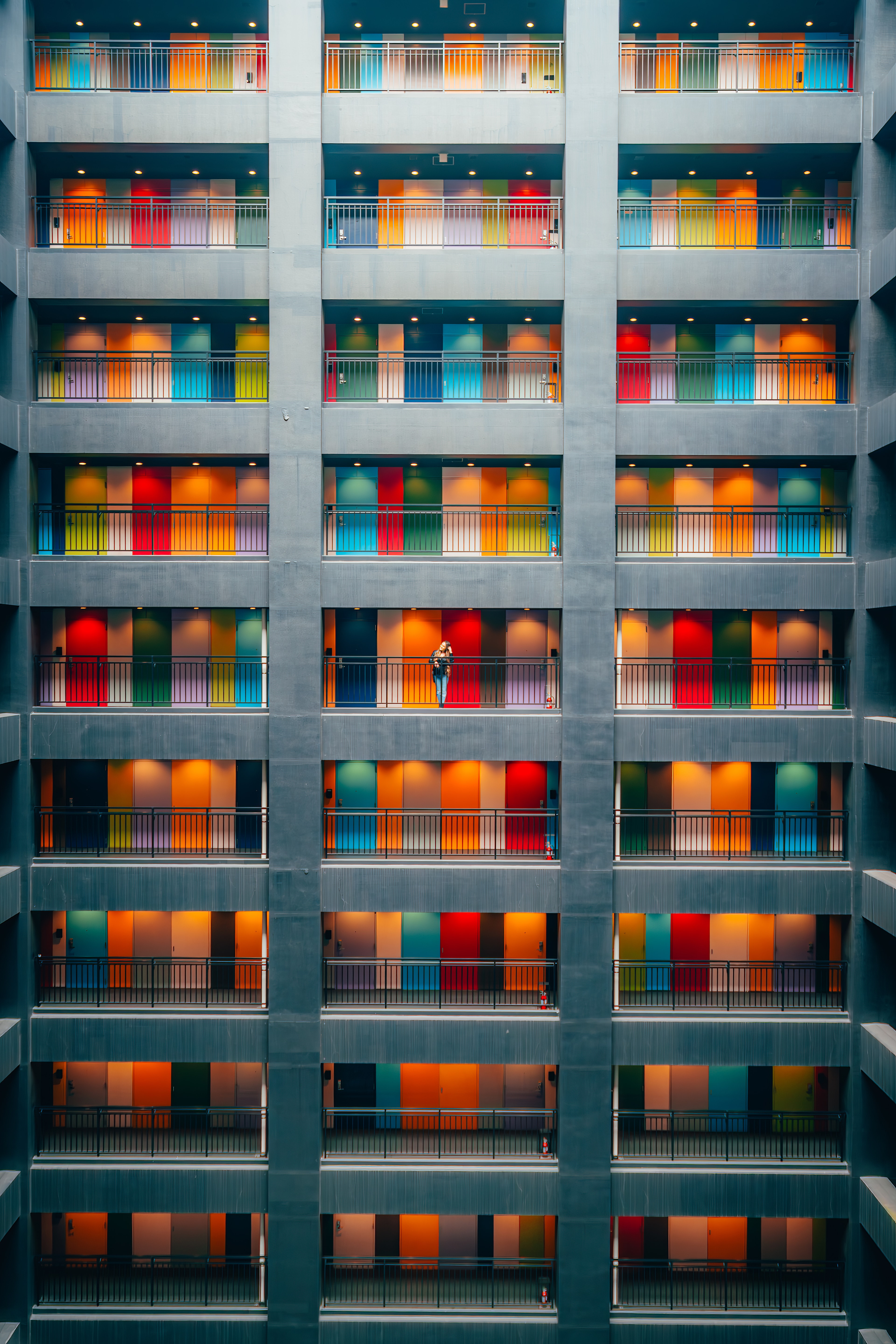 A grid of candy-colored balconies climbing a concrete atrium, one tiny figure leaning out