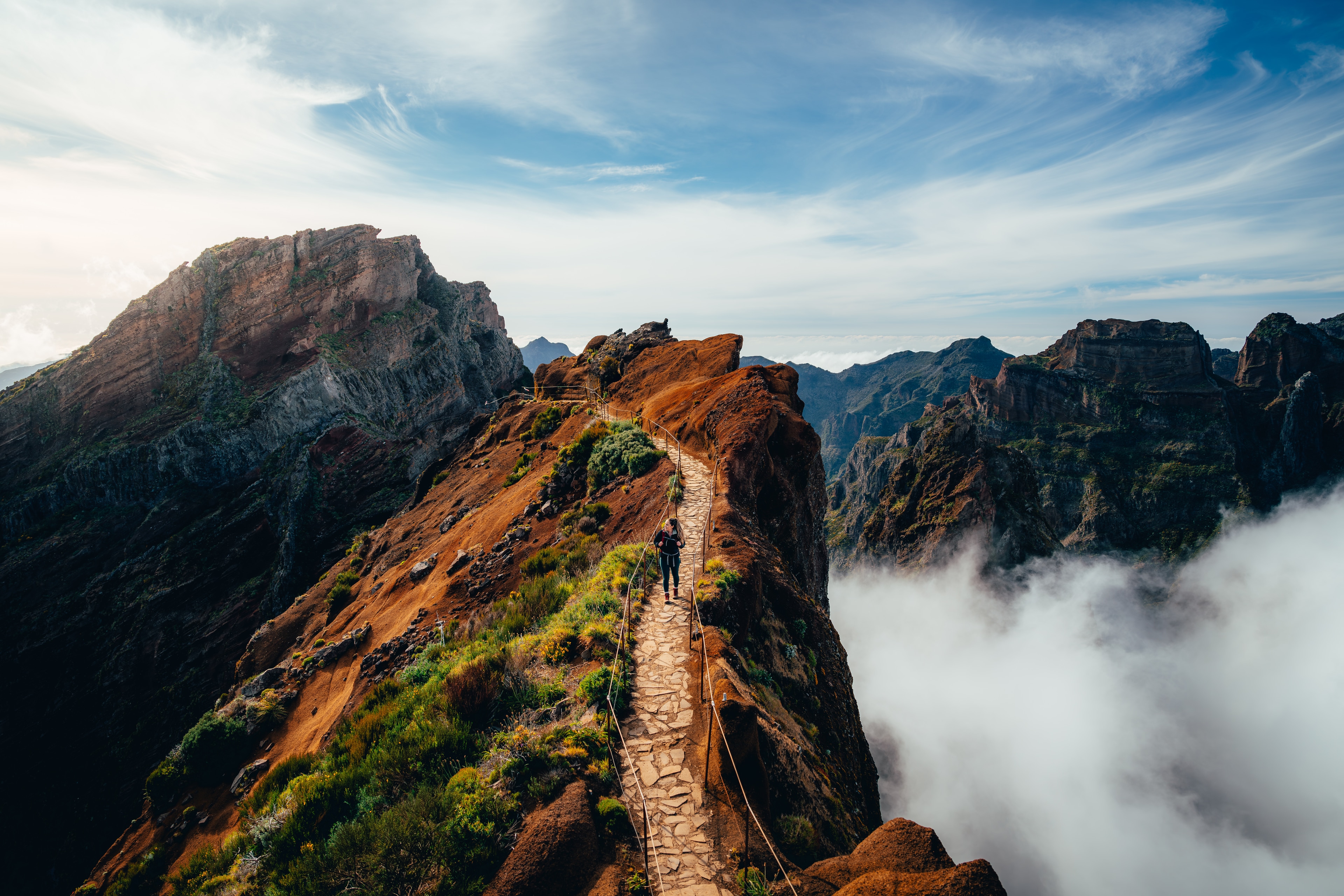 A hiker on a narrow stone ridge path at Pico do Arieiro, peaks rising from a sea of cloud beyond